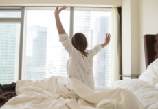 Kvinde sidder i sengen og er klar til en ny dag. Kigger ud på skyskrabere.