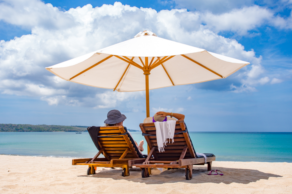 Strand. to personer med ryggen til liggende i solvogne. Stor strandparasol.