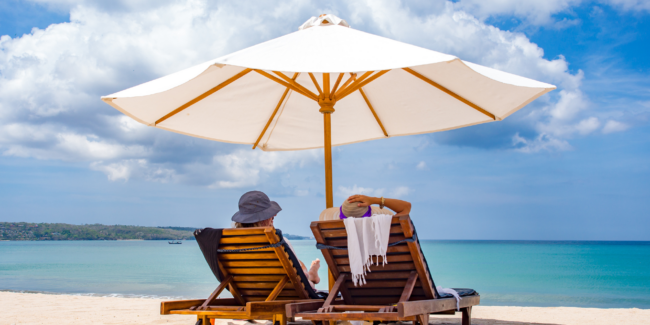 Strand. to personer med ryggen til liggende i solvogne. Stor strandparasol.